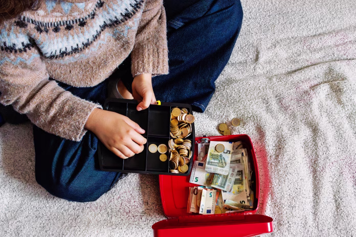 Top view of a young woman sitting on a bed while counting euro coins in an open red cash box filled with euro banknotes