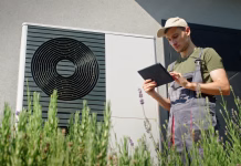 Zakaj je vse več zanimanja za toplotne črpalke? A young technician in overalls and a cap uses a tablet to inspect an outdoor heat pump unit installed on the exterior wall of a building. The scene is set on a sunny day, with the technician surrounded by lavender plants, adding a touch of nature to the industrial setting. The image emphasizes the integration of technology and nature in modern energy solutions.