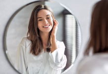 Kako visoko naj bo postavljeno ogledalo v kopalnici? Beauty Concept. Portrait Of Attractive Happy Woman Looking At Mirror In Bathroom, Beautiful Millennial Lady Wearing White Silk Robe Smiling To Reflection, Enjoying Her Appearance, Selective Focus