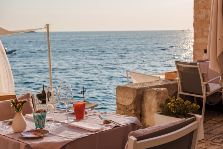 A charming table set for two invites diners to enjoy a sunset meal overlooking the shimmering waters of the Adriatic Sea in Rovinj, Croatia. The atmosphere is warm and serene.