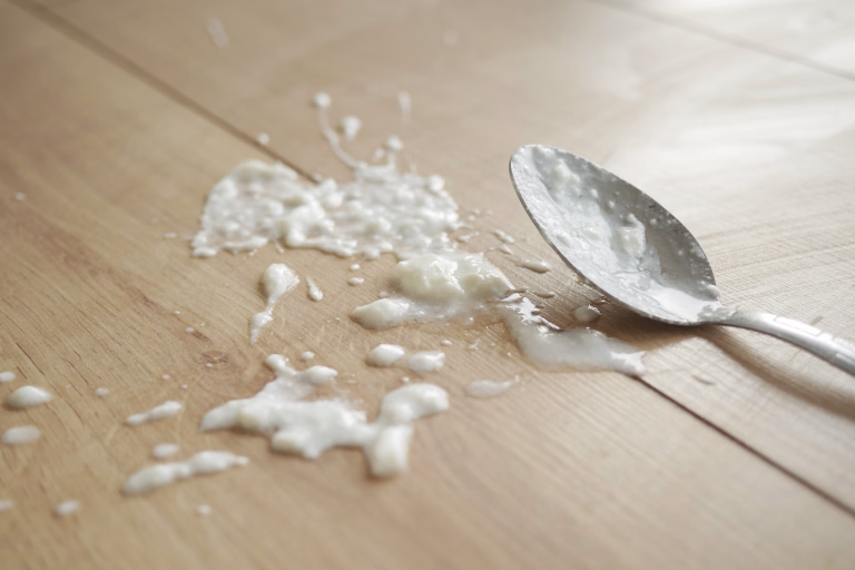 Close-up of spilled yogurt on a wooden floor with a spoon in the foreground. Clean-up concept.