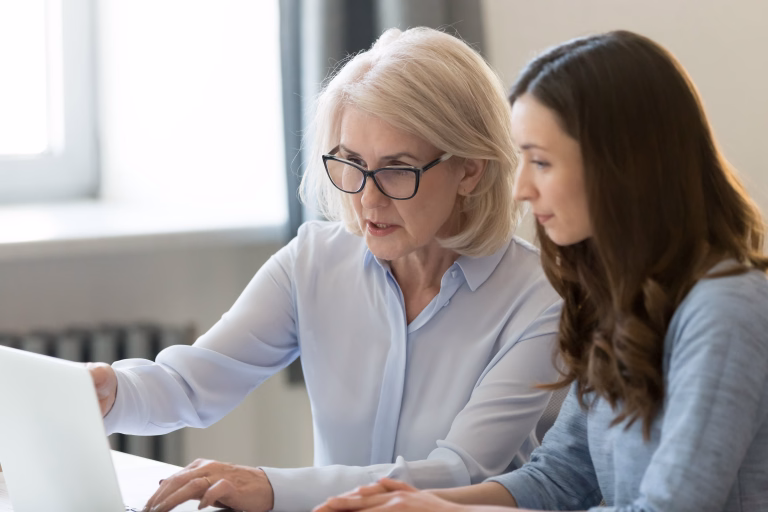 Serious old female mentor teacher coach teaching intern or student computer work pointing at laptop, mature executive manager explaining online project to young employee learning new skills in office