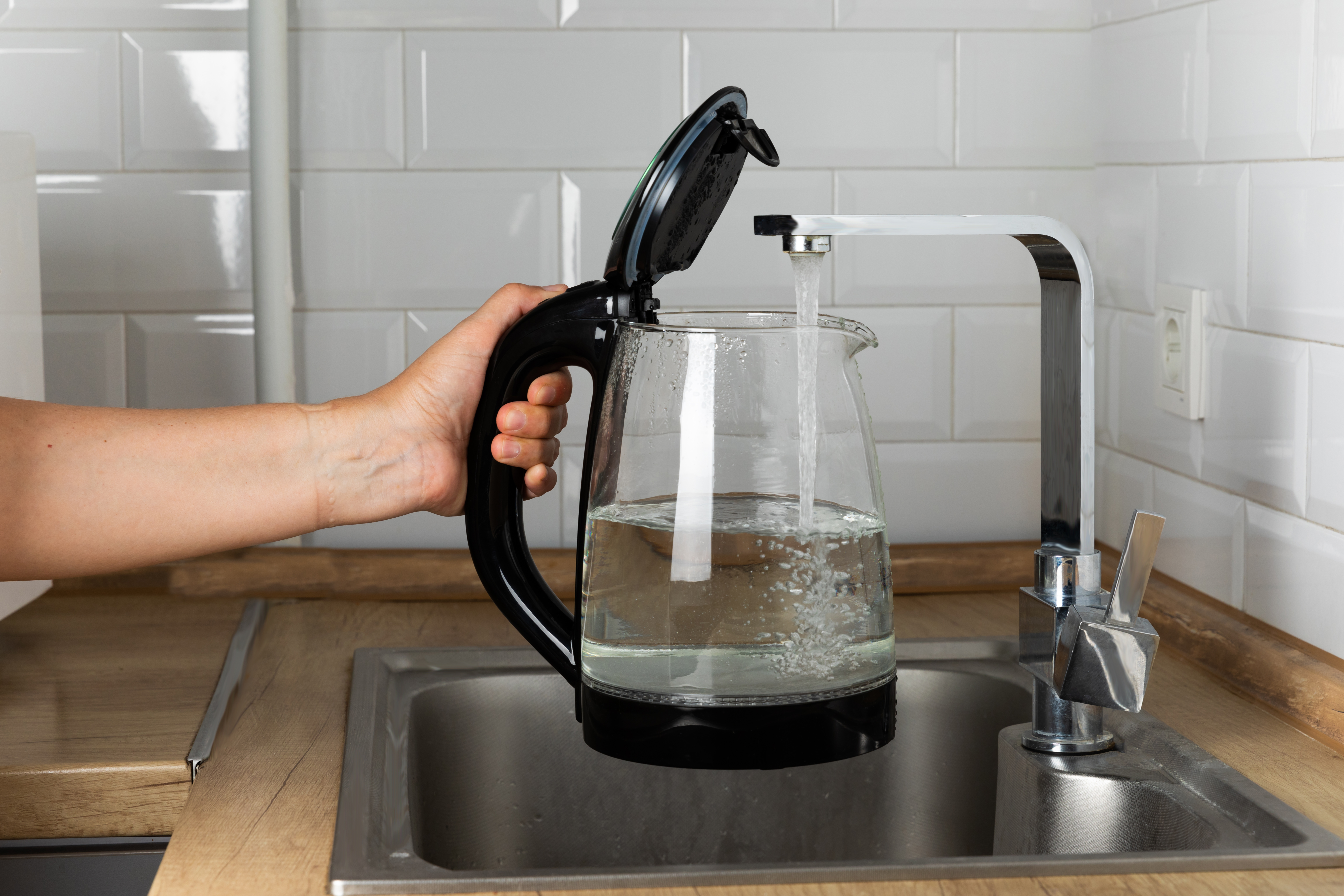 A woman draws water into an electric kettle in the background of the kitchen. It's time for breakfast and tea. Modern electric kettle on a wooden table. Kettle for boiling water.