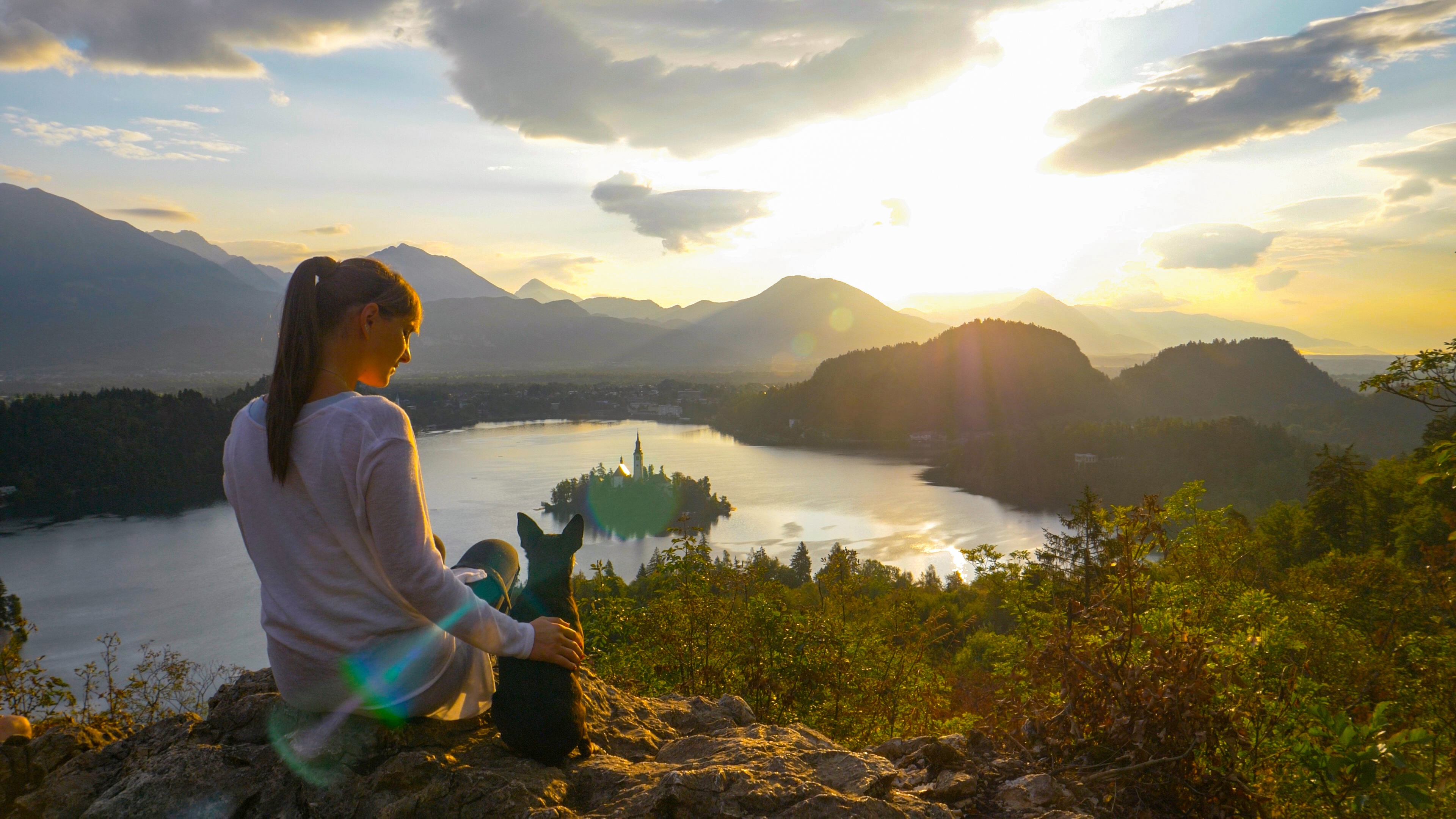 CLOSE UP, LENS FLARE, COPY SPACE: Young Caucasian woman observing the stunning lake below her while petting her adorable dog on a sunny summer evening. Happy female tourist above idyllic lake Bled.