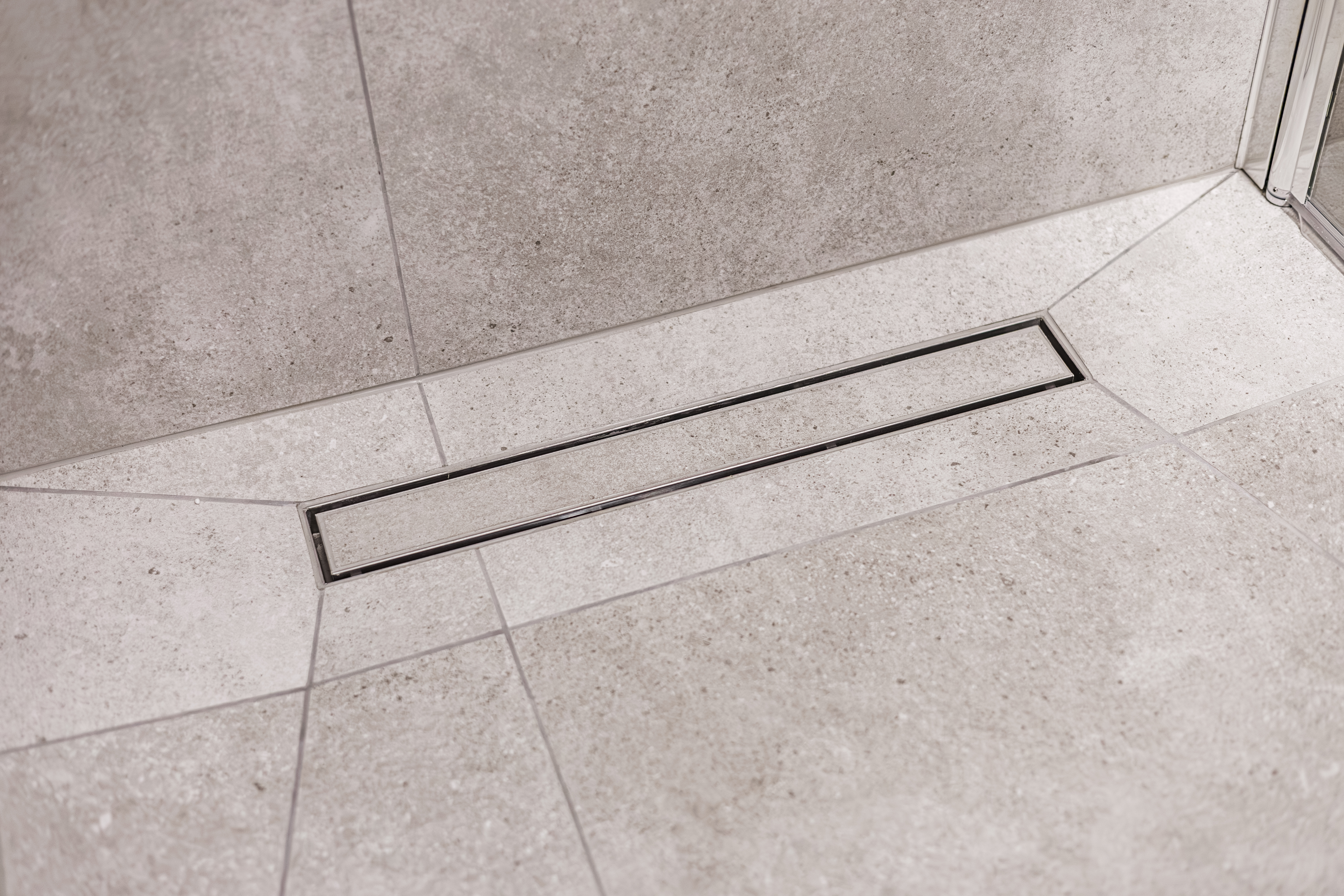 Close-up of a sleek linear drain integrated into a light gray stone-tiled shower floor. Clean, geometric lines emphasize modern bathroom design