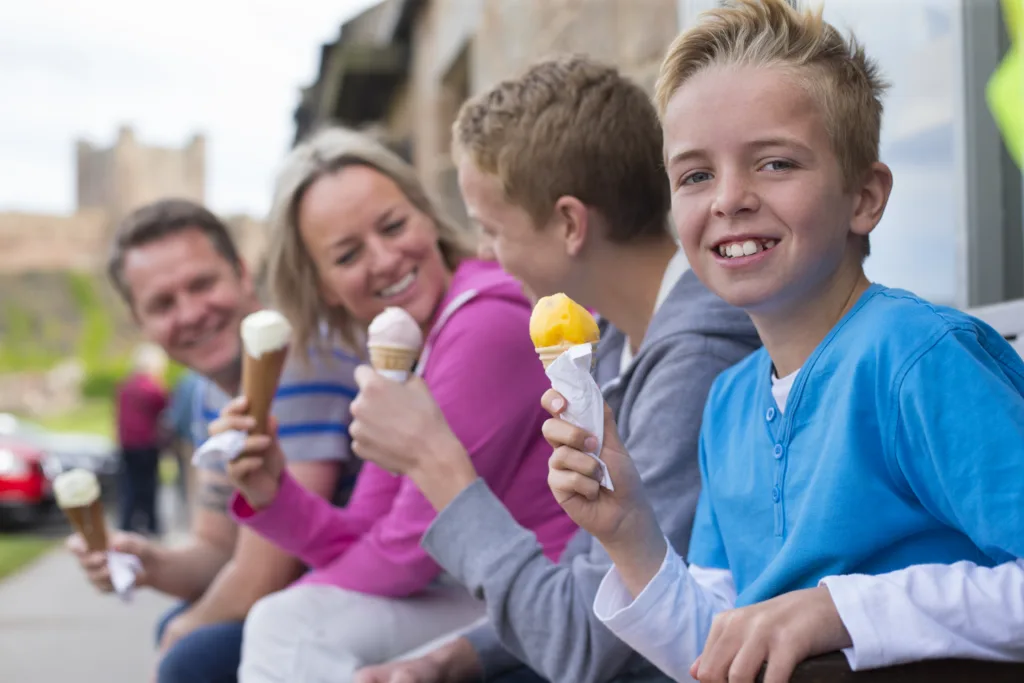 Family enjoying ice cream outside of a shop in a village. They are all holding their cones and a castle can be seen in the background in the distance.