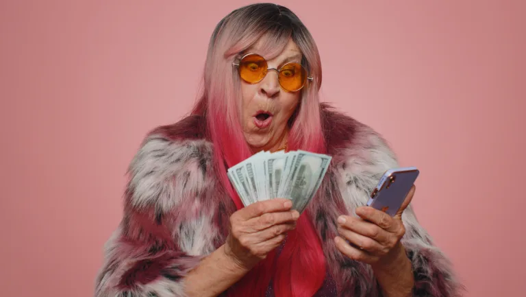 Happy tourist woman looking smartphone display sincerely rejoicing win, receiving money dollar cash banknotes, success lottery luck. Senior stylish grandmother isolated alone on blue studio background