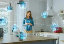 Wi-Fi v hiši: zakaj je planiranje pomembno že med gradnjo Internet of Things Concept: Young Woman Using Smartphone in Kitchen. She controls her Kitchen Appliances with IOT. Graphics Showing Digitalization Visualization of Connected Home Electronics Devices