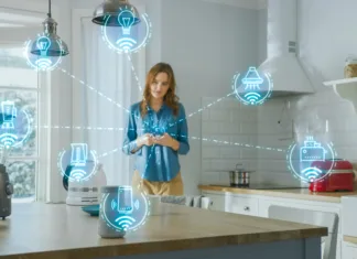 Wi-Fi v hiši: zakaj je planiranje pomembno že med gradnjo Internet of Things Concept: Young Woman Using Smartphone in Kitchen. She controls her Kitchen Appliances with IOT. Graphics Showing Digitalization Visualization of Connected Home Electronics Devices