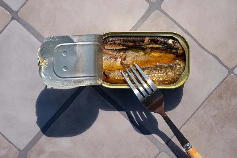 Overhead view of open sardine tin with a fork inside, casting long shadows on a tiled surface. Simple food or emergency meal.