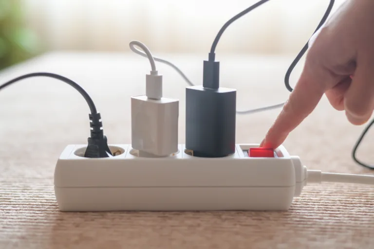 Person pressing red on/off switch on white power strip to cut electricity to multiple chargers, symbolizing mindful power use, energy saving, lower bills and household sustainability