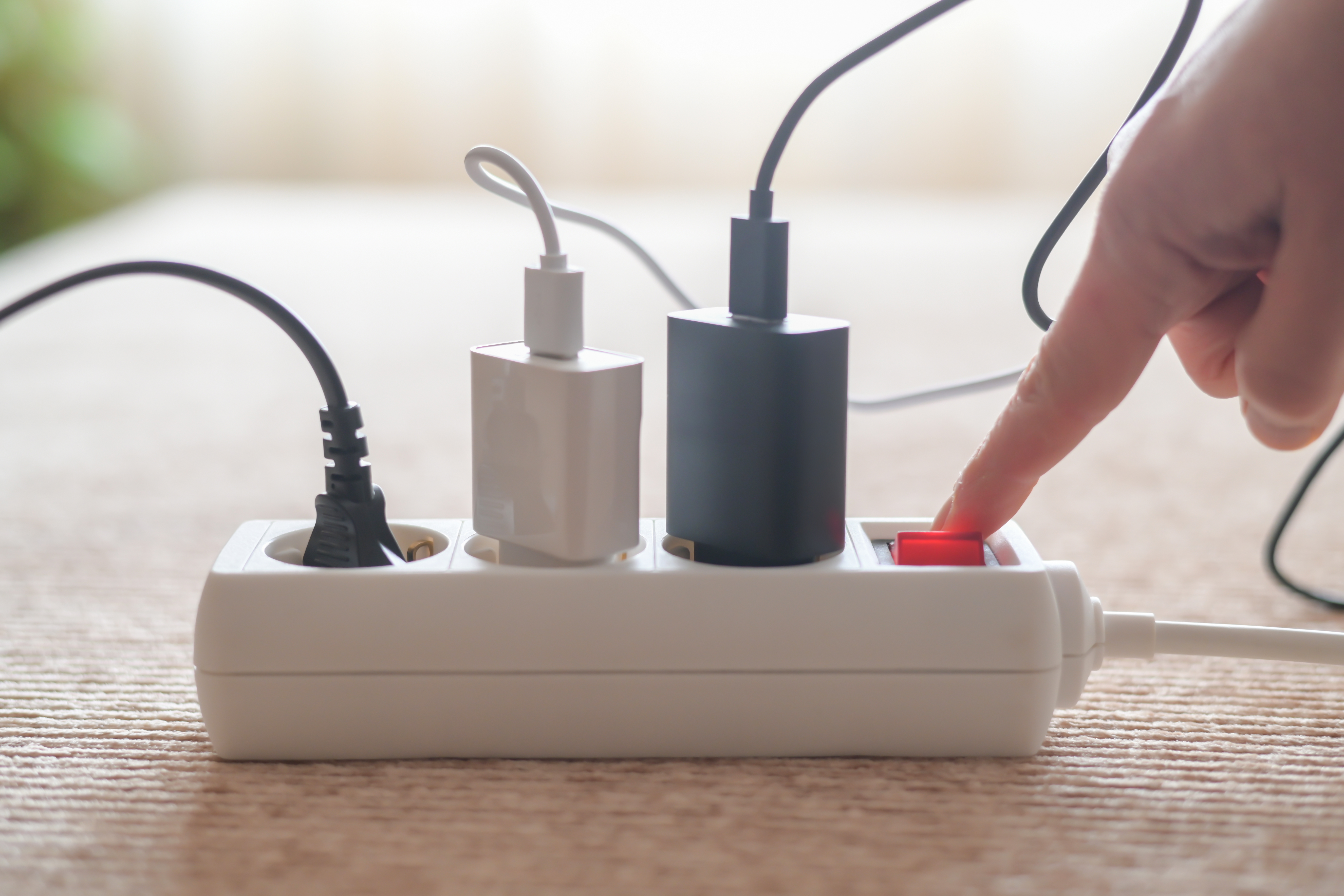 Ta naprava porabi več elektrike kot klimatska naprava, izklopi pa jo le malokdo: to najbolj vpliva na račun za elektriko Person pressing red on/off switch on white power strip to cut electricity to multiple chargers, symbolizing mindful power use, energy saving, lower bills and household sustainability
