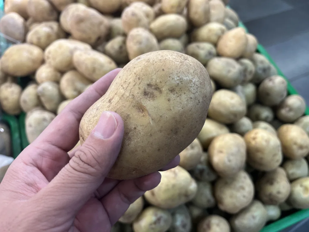 A close-up shot shows a hand holding an earthy brown potato over a green bin filled with more fresh potatoes. Ideal for grocery shopping blogs or food nutrition guides.