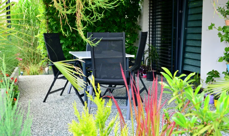 Beautiful outdoor house patio with stone carpet flooring, growing green flowers and plants, and table with chairs. House terrace overgrown with plants with garden furniture.