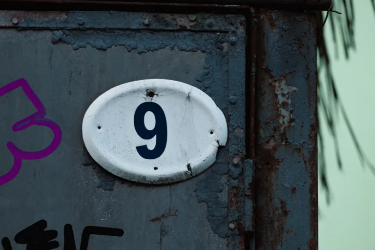 Close-up of house number 9 on worn metal surface with rust, cracks and peeling paint. Urban grunge texture with industrial character, conveying age, decay and raw architectural detail.