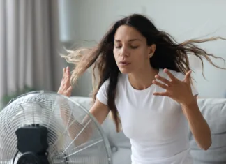 Strokovnjaki opozarjajo: ventilatorja nikar ne uporabljajte, ko temperature presežejo to mejo Millennial Caucasian girl sit on sofa in living room breathe fresh air from floor ventilator suffer from summer heat at home, young woman feel hot use fan at home struggle with lack of air condition