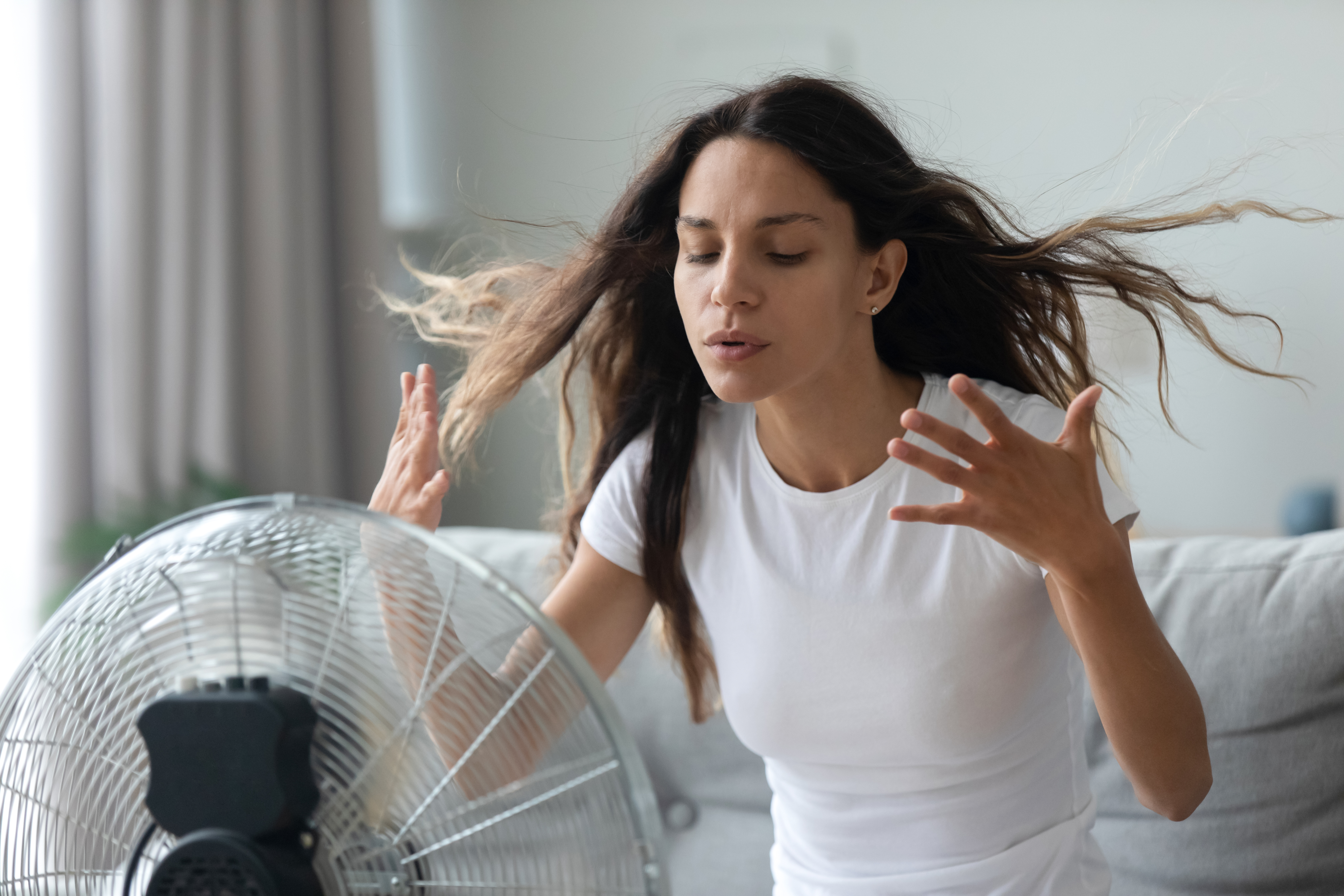 Strokovnjaki opozarjajo: ventilatorja nikar ne uporabljajte, ko temperature presežejo to mejo Millennial Caucasian girl sit on sofa in living room breathe fresh air from floor ventilator suffer from summer heat at home, young woman feel hot use fan at home struggle with lack of air condition