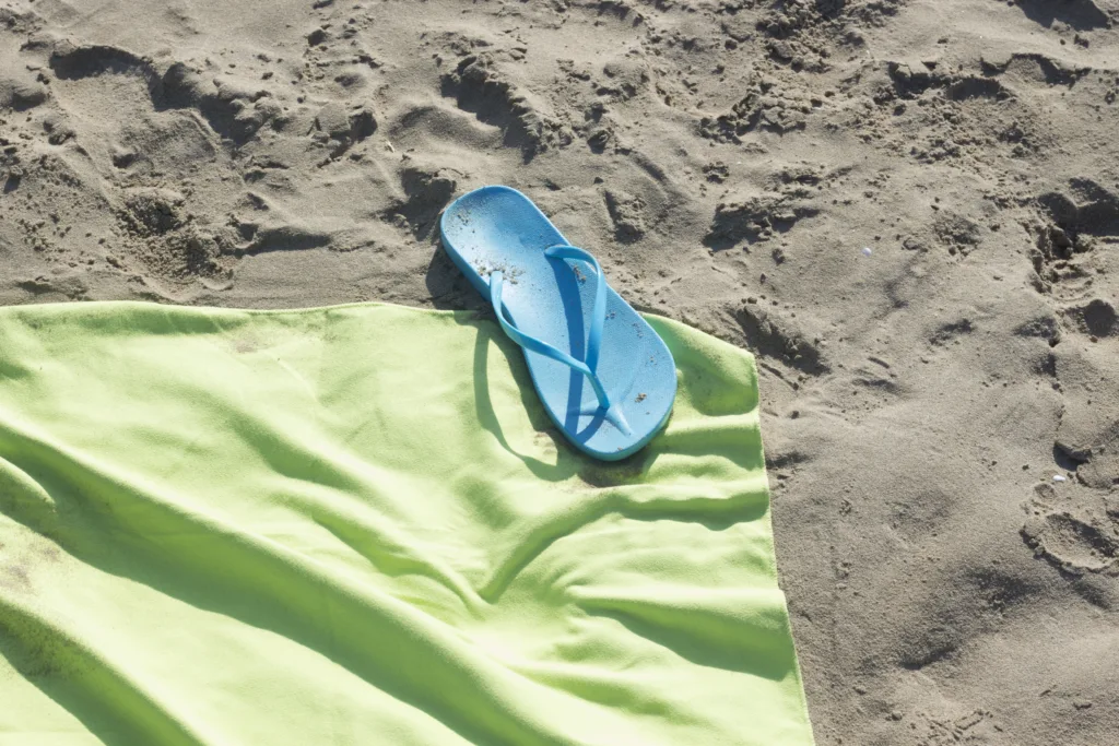 Towel and sandals on sandy beach by the ocean on hot summer day.