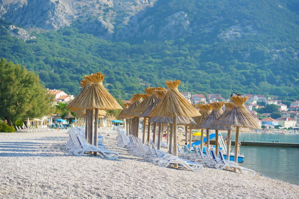 Traditional straw parasols lined up along the famous pebble beach in Baška, with lush green hills and rocky mountains rising above the Adriatic coast. Ideal concept for sustainable tourism, beach holidays and Mediterranean lifestyle