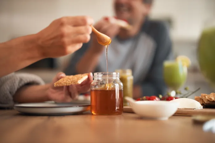Two people enjoy a cozy breakfast at home, dipping bread into honey from a jar. A warm, inviting scene of togetherness with spreads, fruit, and juice.
