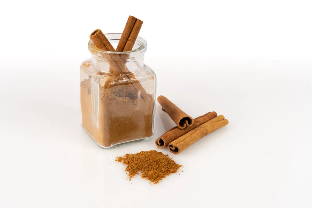 A vibrant studio shot features aromatic cinnamon in various forms on a clean white background. A clear, square glass jar is filled with fine ground cinnamon powder, with two whole cinnamon sticks elegantly placed inside. Adjacent to the jar, three additional natural cinnamon quills are neatly arranged alongside a small, inviting pile of the powdered spice. The bright, even lighting accentuates the rich, warm brown hues and rustic texture of the spice, making it ideal for culinary, health, or lifestyle projects requiring a fresh, natural ingredient focus.