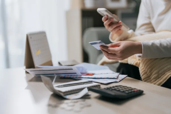 Woman holding credit card and smartphone with unpaid bills on table, representing household debt and extravagant spending lifestyle.