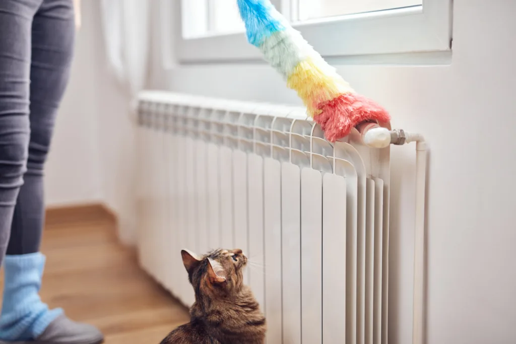 Woman with a dust stick cleaning central heating gas radiator at home.